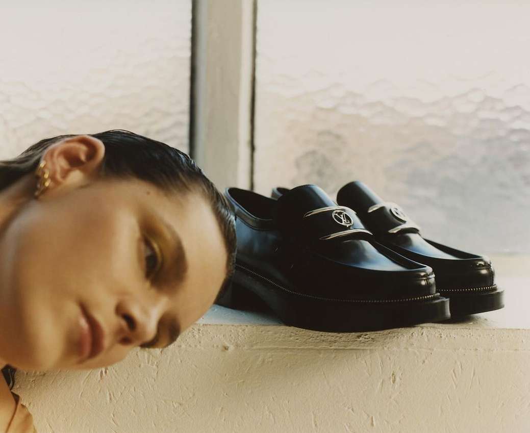 Close-up of a woman with her head resting near black leather penny loafer shoes with silver hardware, positioned by a bright window.