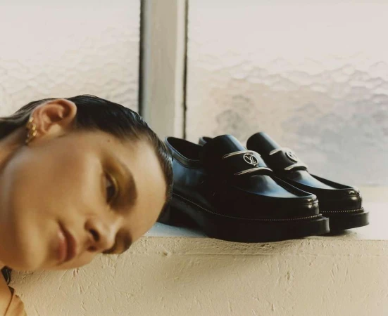 Close-up of a woman with her head resting near black leather penny loafer shoes with silver hardware, positioned by a bright window.