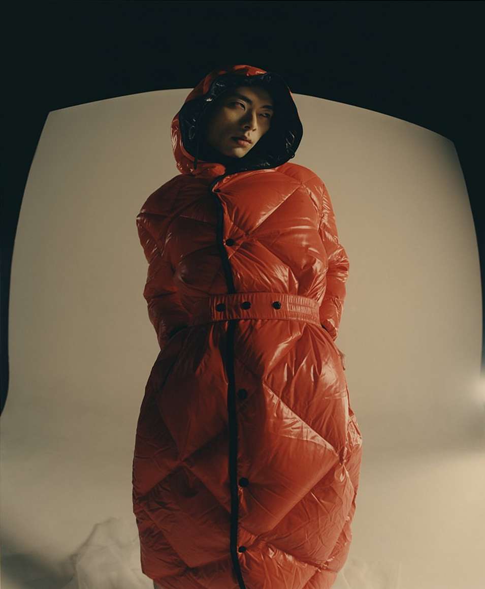 A person in a shiny red puffer coat with a hood gazes slightly left against a soft studio backdrop with subtle lighting and visual depth.