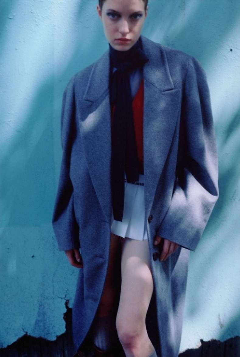 Fashion image featuring a stylish adult female model in a gray coat, black turtleneck, and white skirt against a textured blue wall.