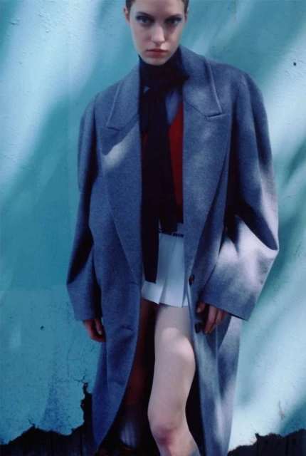 Fashion image featuring a stylish adult female model in a gray coat, black turtleneck, and white skirt against a textured blue wall.