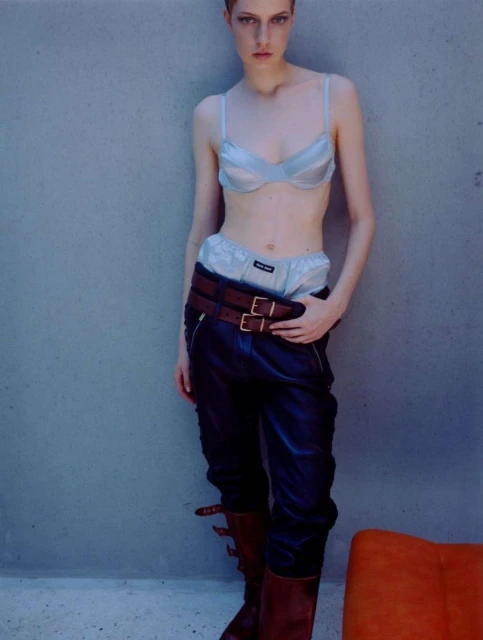 A slender woman with short hair poses in a light blue bra, briefs, and shiny dark leather pants, paired with high brown leather boots.