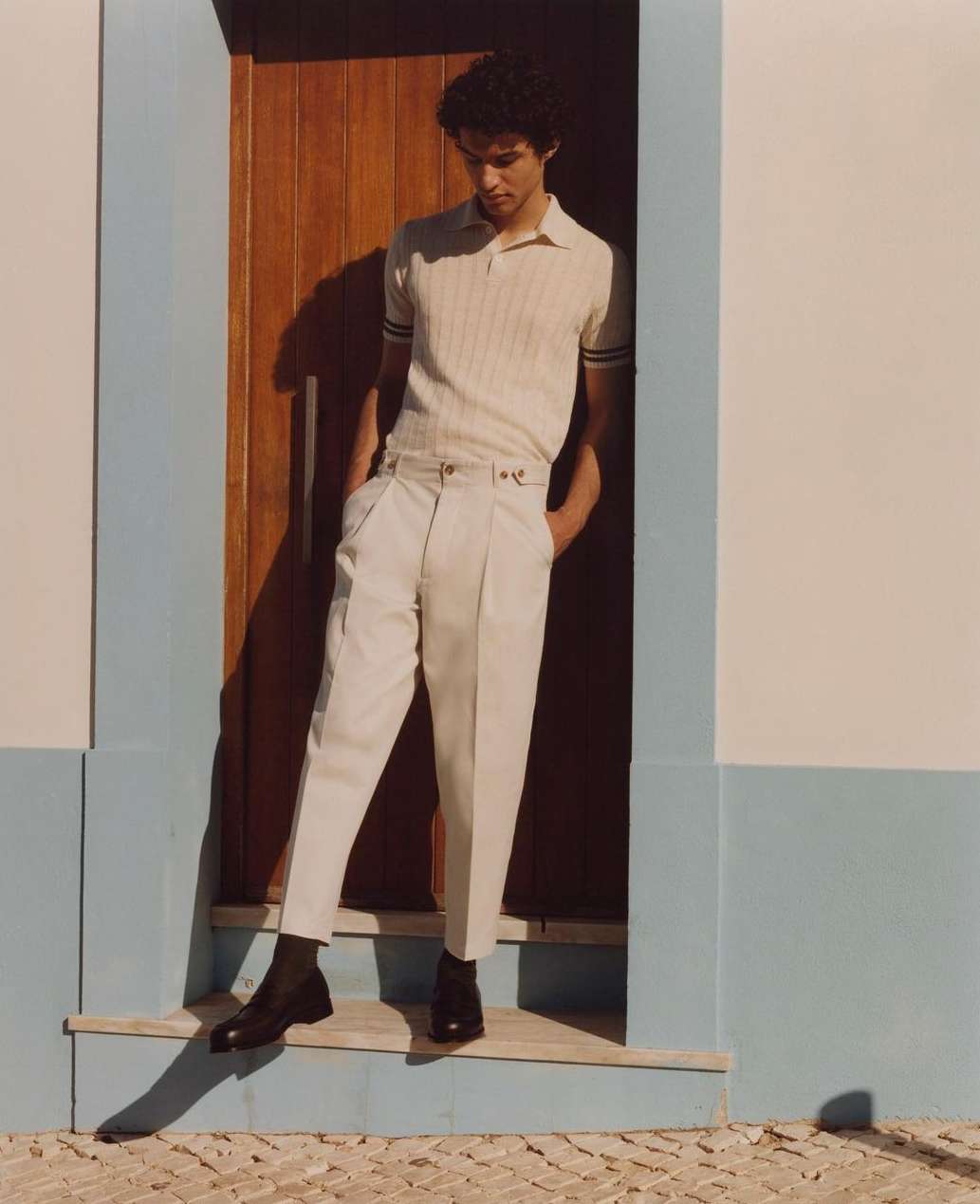 A young man in a tan polo shirt and cream-colored pants poses in a doorway. He is standing on a doorstep in black shoes.