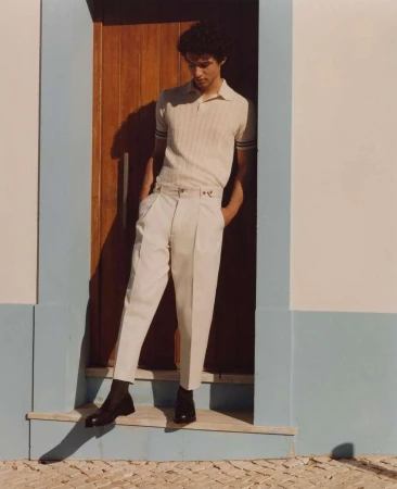 A young man in a tan polo shirt and cream-colored pants poses in a doorway. He is standing on a doorstep in black shoes.