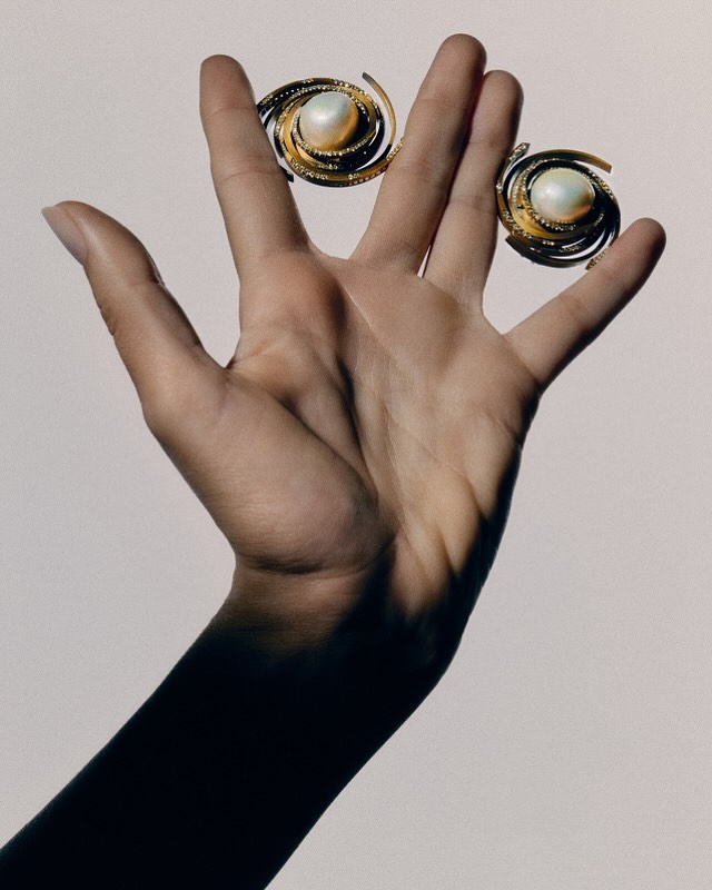 Hand holding two gold spiral earrings with pearl centers against a light background in a close-up studio product shot.