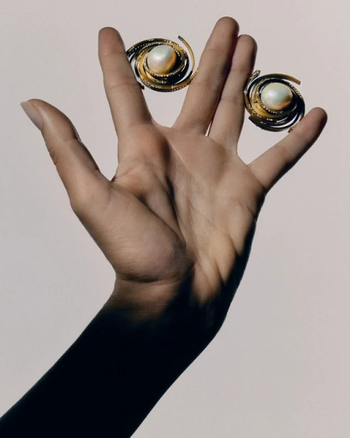 Hand holding two gold spiral earrings with pearl centers against a light background in a close-up studio product shot.