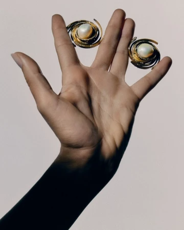 Hand holding two gold spiral earrings with pearl centers against a light background in a close-up studio product shot.