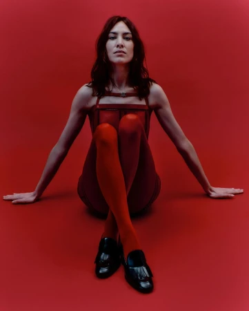 Brunette woman in red dress, red stockings, and black loafers poses against a red background with her arms outstretched on the floor.