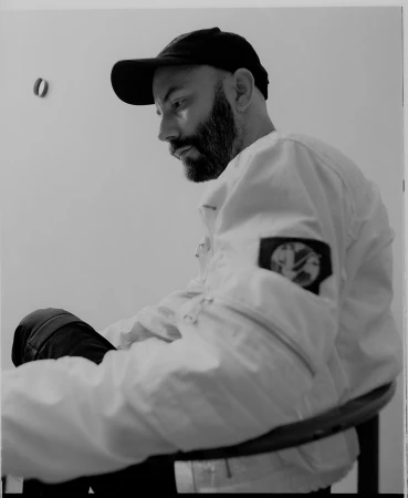 Man wearing a baseball cap and light jacket seated in a chair against a plain white wall