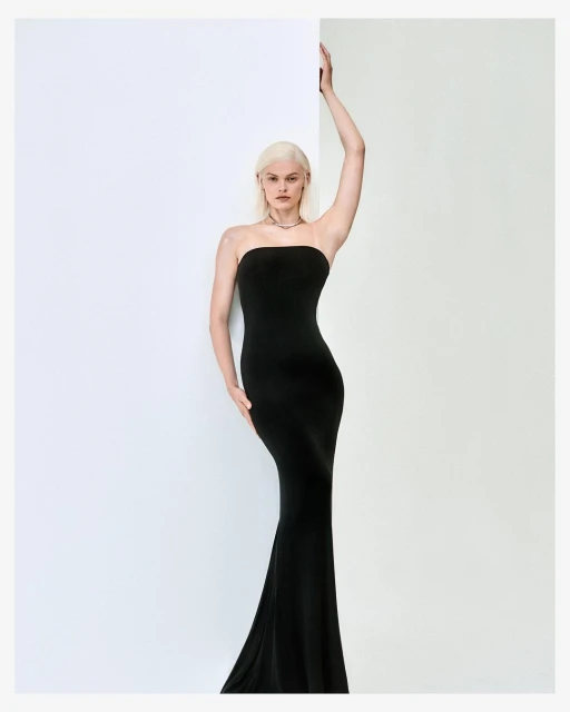 A woman posing in a black strapless evening gown stands against a split white and light gray background, showcasing fashion and elegance.