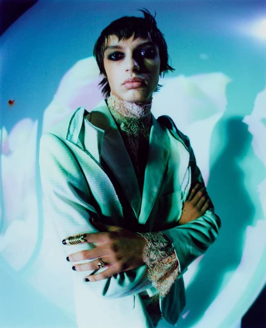 Portrait of an adult male with dark hair wearing a light-green blazer, lace collar, and rings with a blurred floral background.