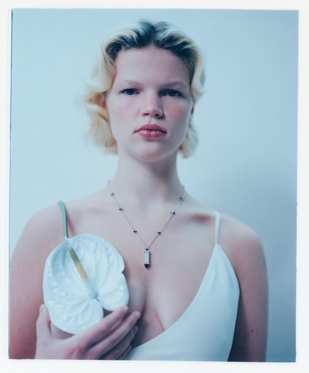 Portrait of a young woman in a white tank top holding a white Anthurium flower against a pale background in a studio setting.