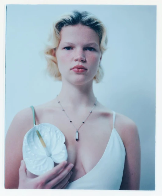 Portrait of a young woman in a white tank top holding a white Anthurium flower against a pale background in a studio setting.
