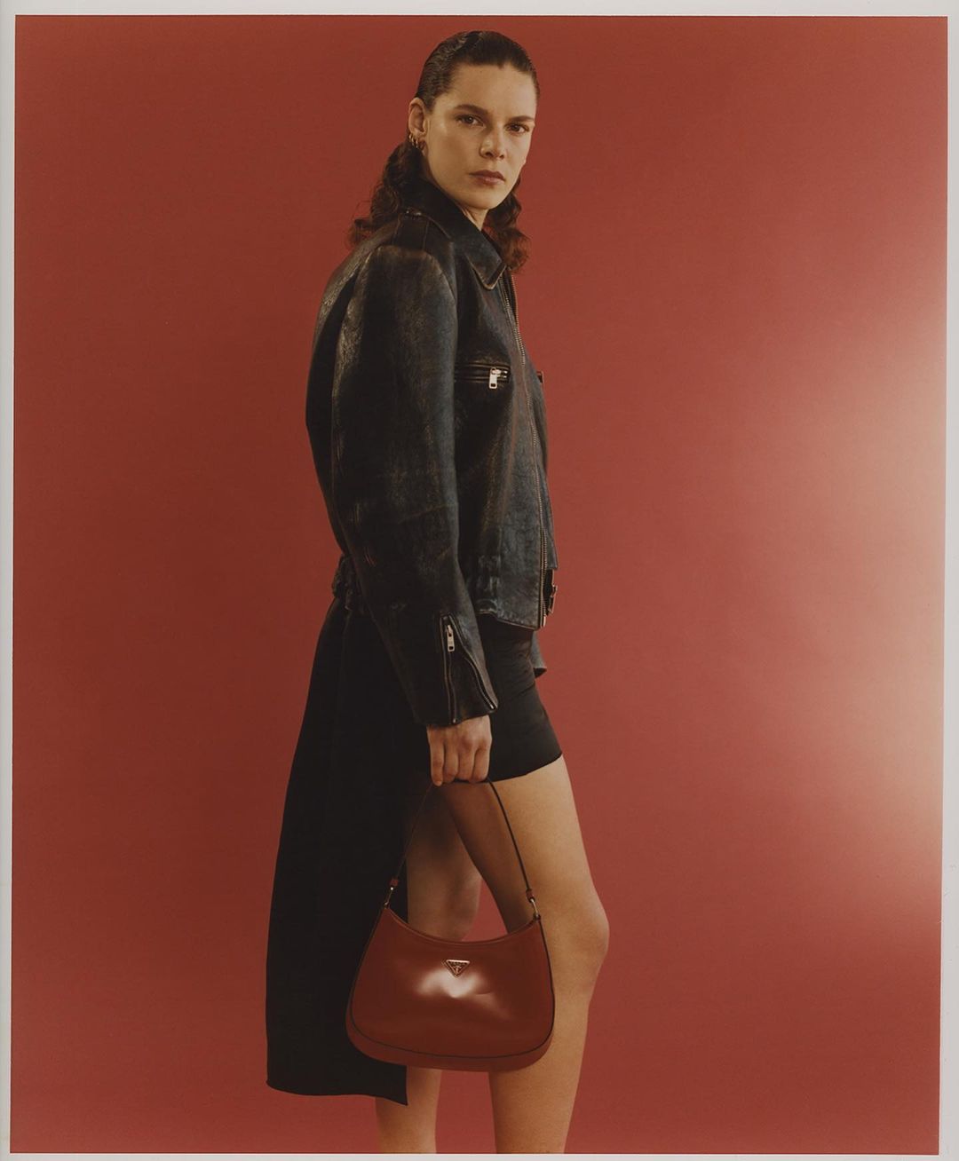 Fashion editorial shot of a woman wearing a black leather jacket and holding a brown leather handbag against a red background.