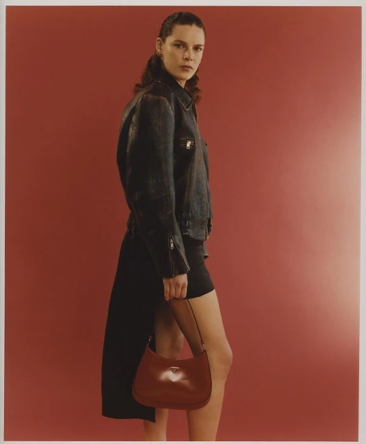 Fashion editorial shot of a woman wearing a black leather jacket and holding a brown leather handbag against a red background.