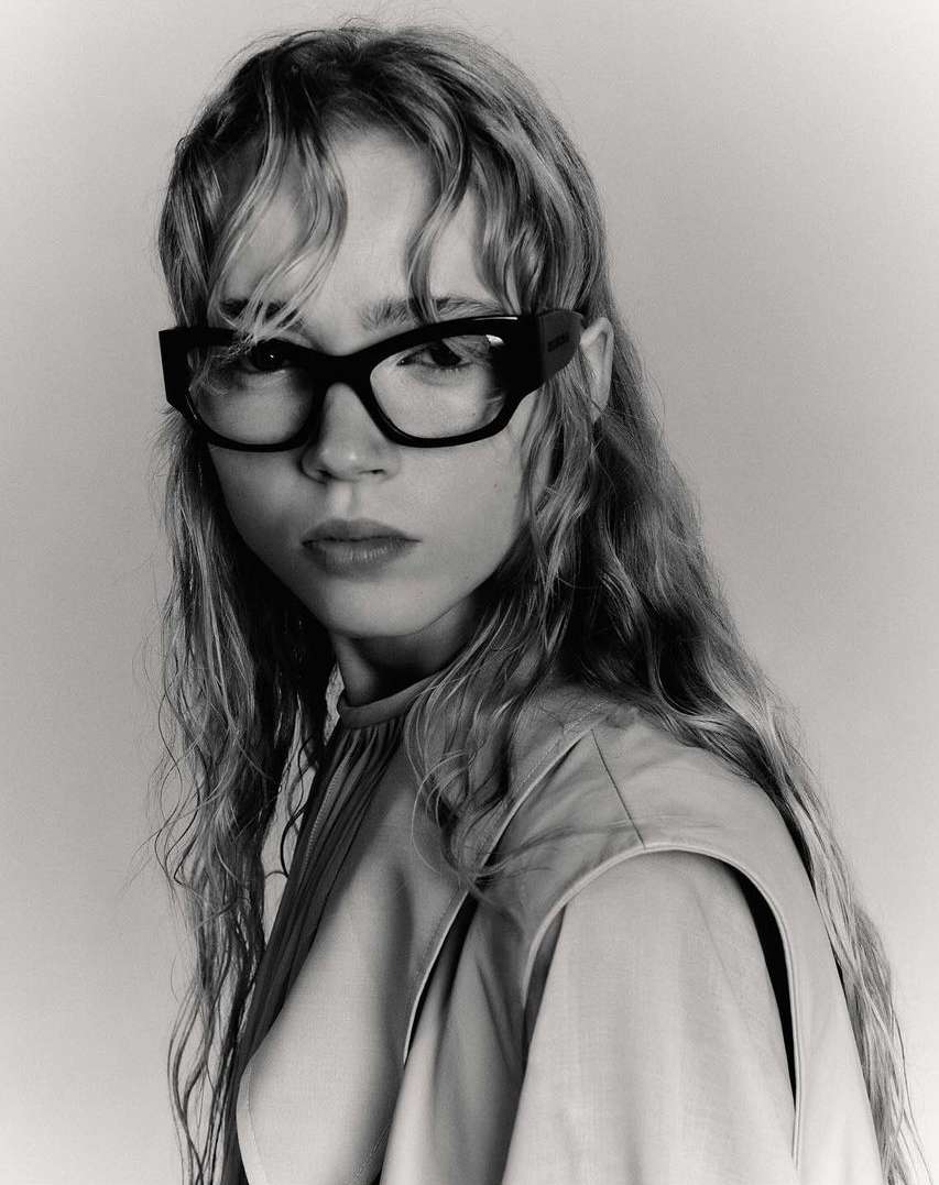 Portrait of a young woman with long wavy hair wearing black-framed glasses and a beige top, captured in a black and white studio shot.