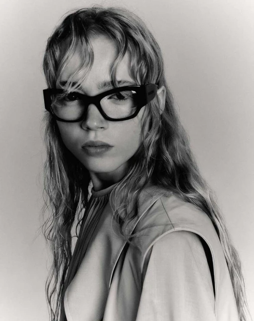Portrait of a young woman with long wavy hair wearing black-framed glasses and a beige top, captured in a black and white studio shot.