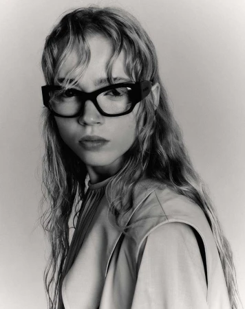 Portrait of a young woman with long wavy hair wearing black-framed glasses and a beige top, captured in a black and white studio shot.
