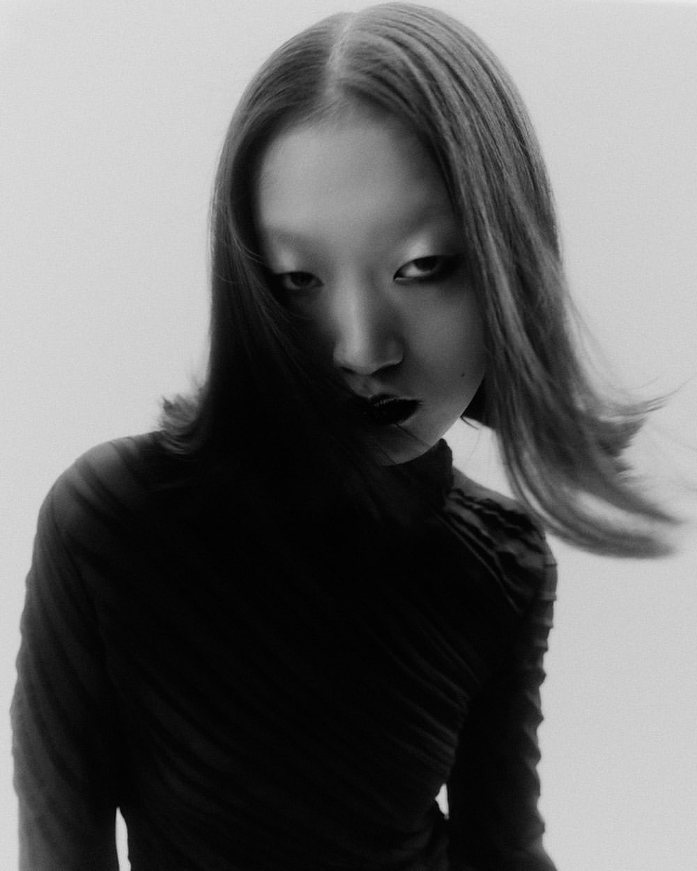 Monochrome portrait of a young Asian woman with dark hair and black lipstick wearing a black turtleneck against a white background.