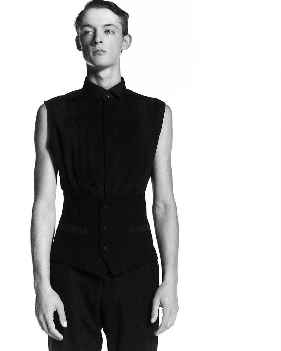 Portrait of a young adult male in a black sleeveless collared shirt and black pants, standing in front of a bright white background.