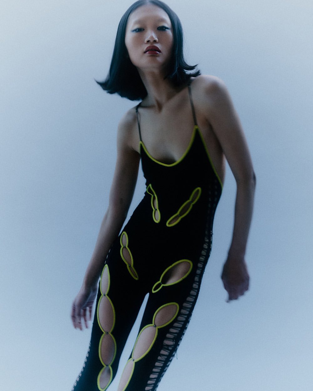 Asian woman posing in black cutout bodysuit against a blue background in a fashion editorial studio shot. High fashion. 
