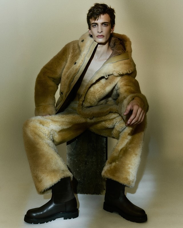 Male model sits wearing a fur-like coat and pants with black boots in a neutral studio setting, posing for a fashion editorial.