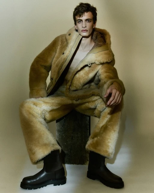 Male model sits wearing a fur-like coat and pants with black boots in a neutral studio setting, posing for a fashion editorial.