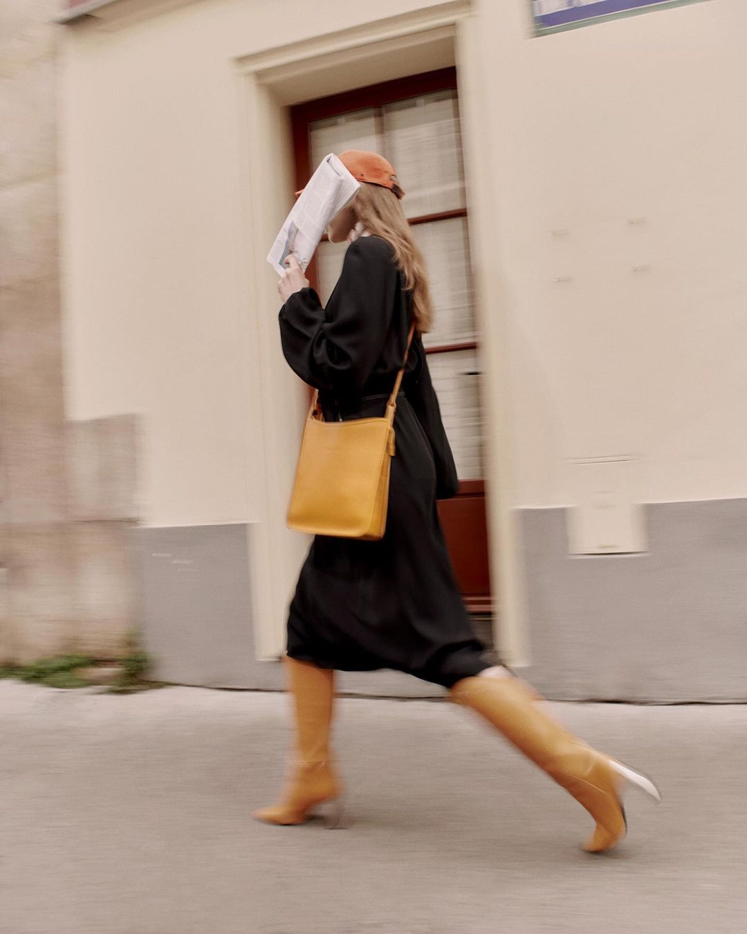 A woman in a black dress walks along a street with a newspaper covering her face, wearing a cap and boots, with a yellow bag.