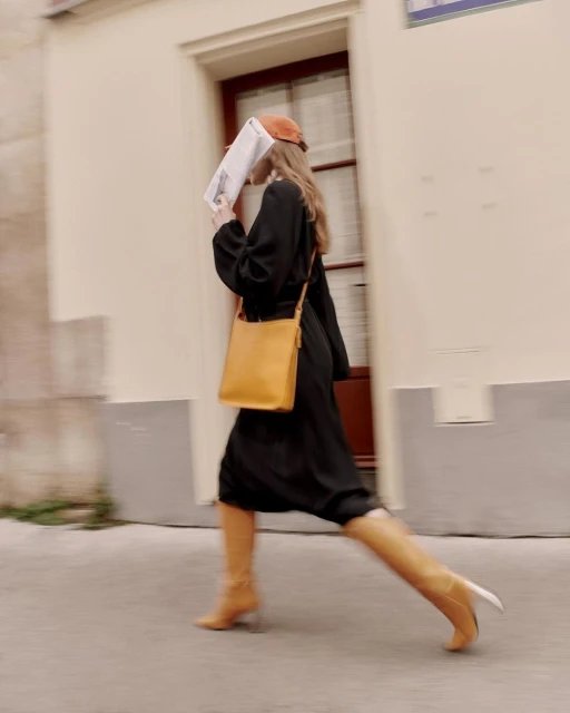 A woman in a black dress walks along a street with a newspaper covering her face, wearing a cap and boots, with a yellow bag.