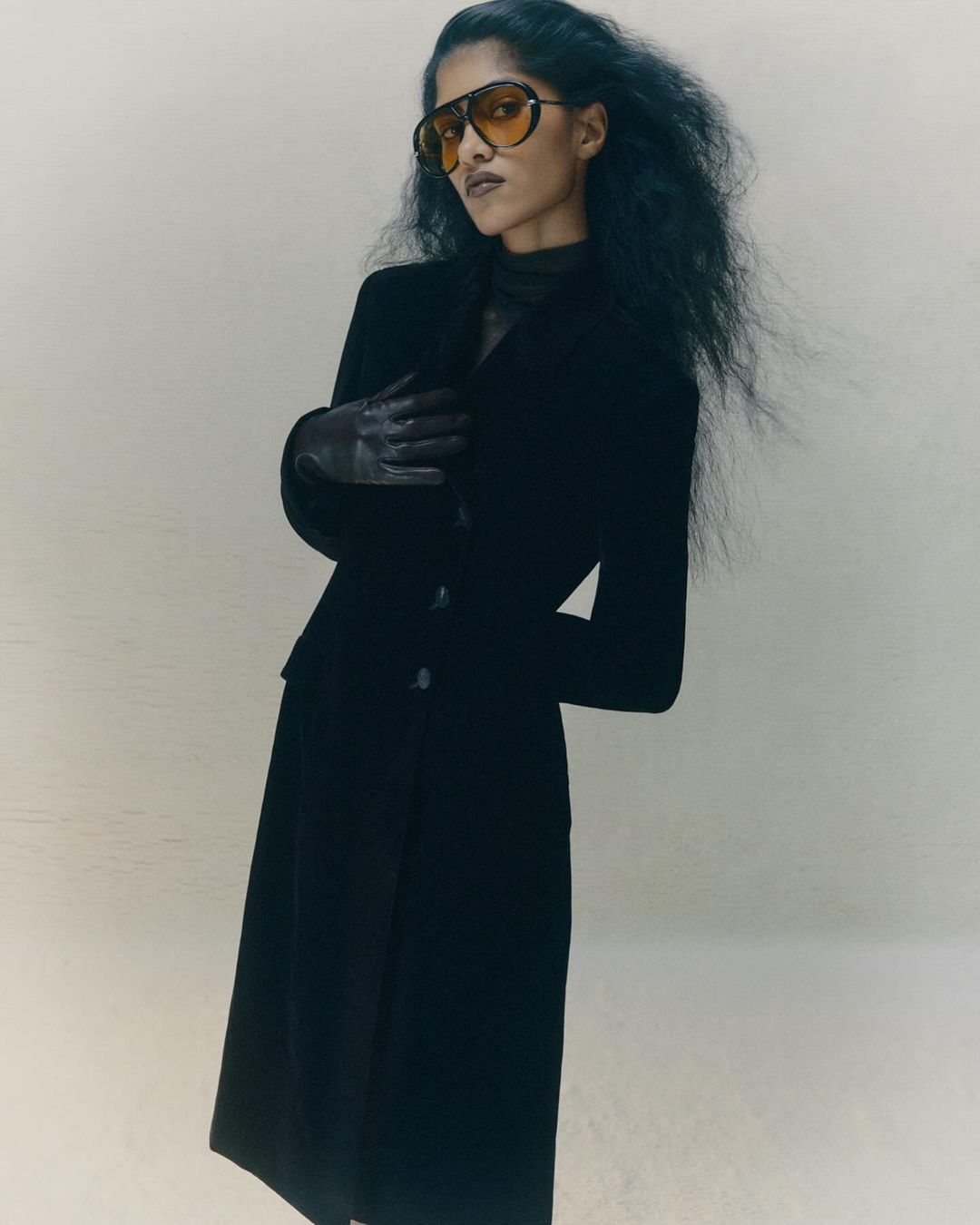 Fashionable woman in black jacket and skirt wearing aviator sunglasses stands in neutral studio with long dark hair and gloved hands.