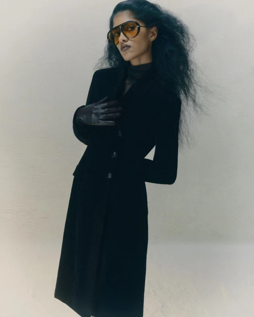 Fashionable woman in black jacket and skirt wearing aviator sunglasses stands in neutral studio with long dark hair and gloved hands.