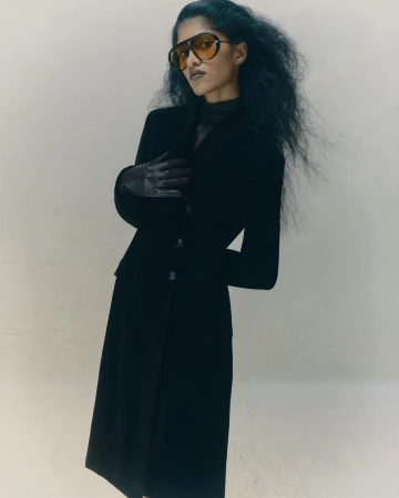 Fashionable woman in black jacket and skirt wearing aviator sunglasses stands in neutral studio with long dark hair and gloved hands.