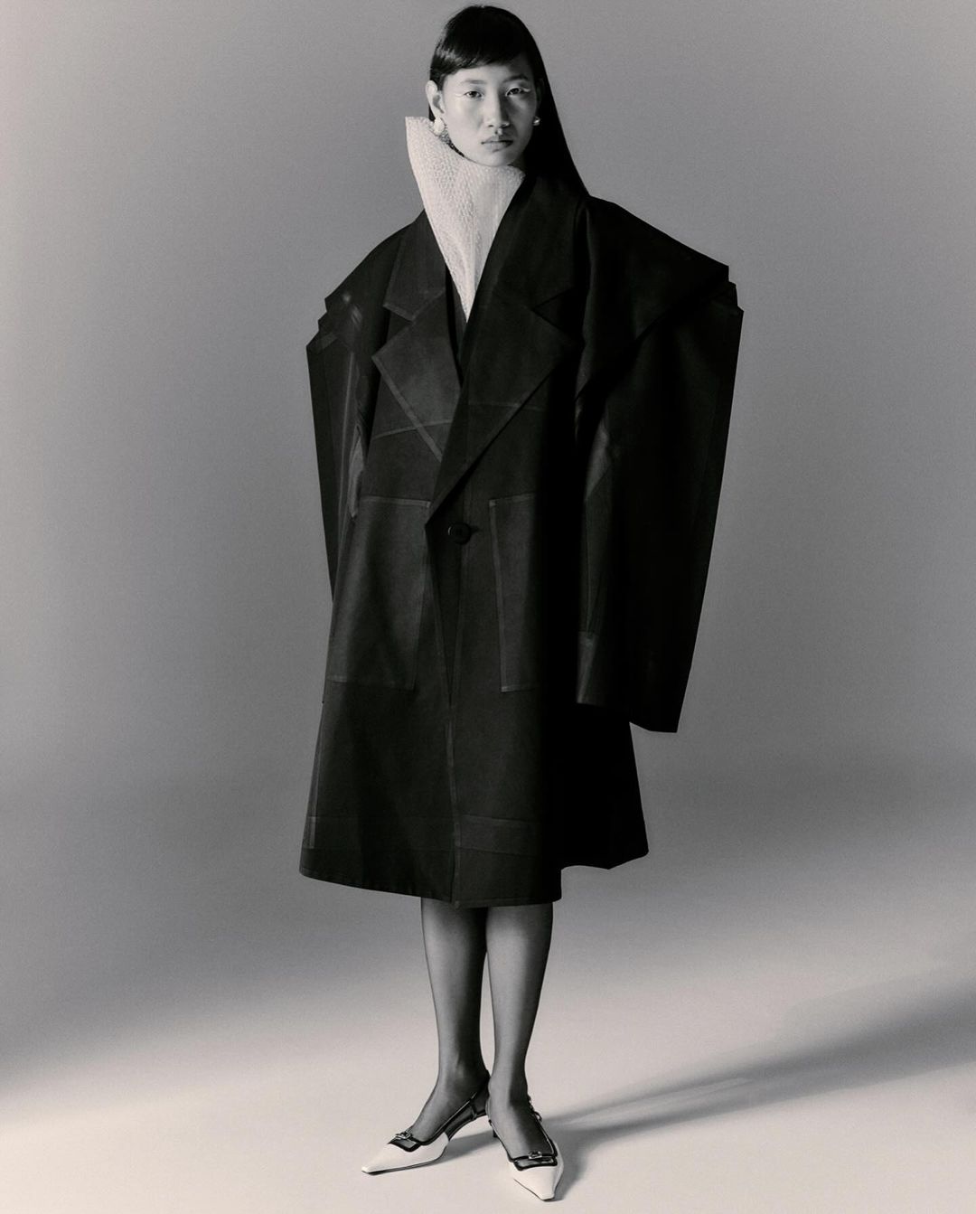 Asian woman in oversized black coat and white mules poses in studio. Tights and white turtleneck. Fashion editorial photography.