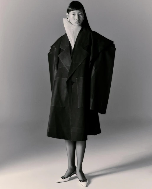 Asian woman in oversized black coat and white mules poses in studio. Tights and white turtleneck. Fashion editorial photography.
