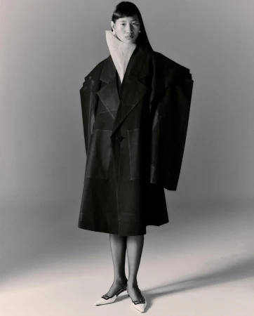 Asian woman in oversized black coat and white mules poses in studio. Tights and white turtleneck. Fashion editorial photography.