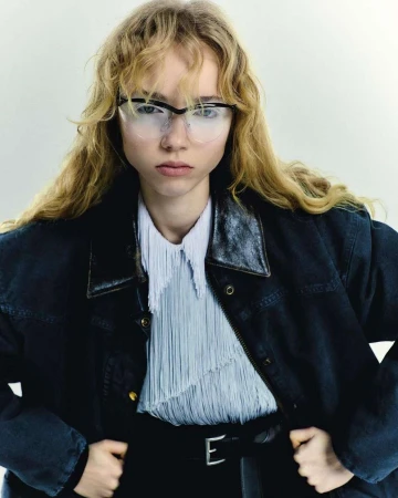 Close-up portrait of a blonde woman wearing glasses, a fringed white shirt, and a dark jacket against a plain white background.