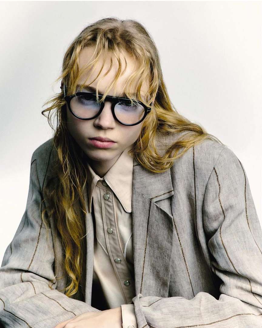 Close-up of a fair-skinned young adult with round sunglasses, a tan button-down shirt, and a grey pinstripe blazer against a white backdrop.