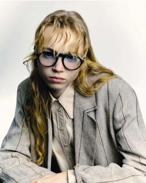 Close-up of a fair-skinned young adult with round sunglasses, a tan button-down shirt, and a grey pinstripe blazer against a white backdrop.