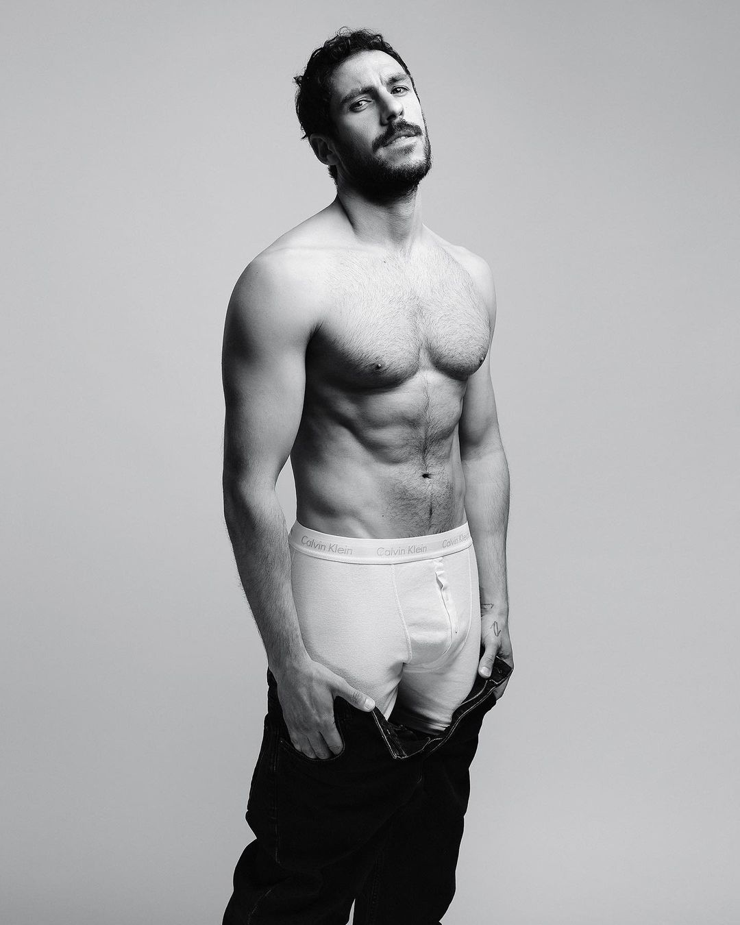 Shirtless man with a beard wearing white briefs and dark unbuttoned trousers against a plain grey background.