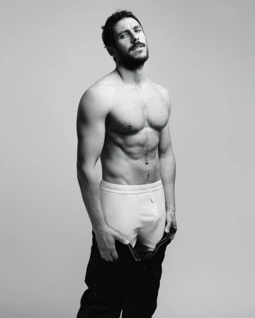 Shirtless man with a beard wearing white briefs and dark unbuttoned trousers against a plain grey background.
