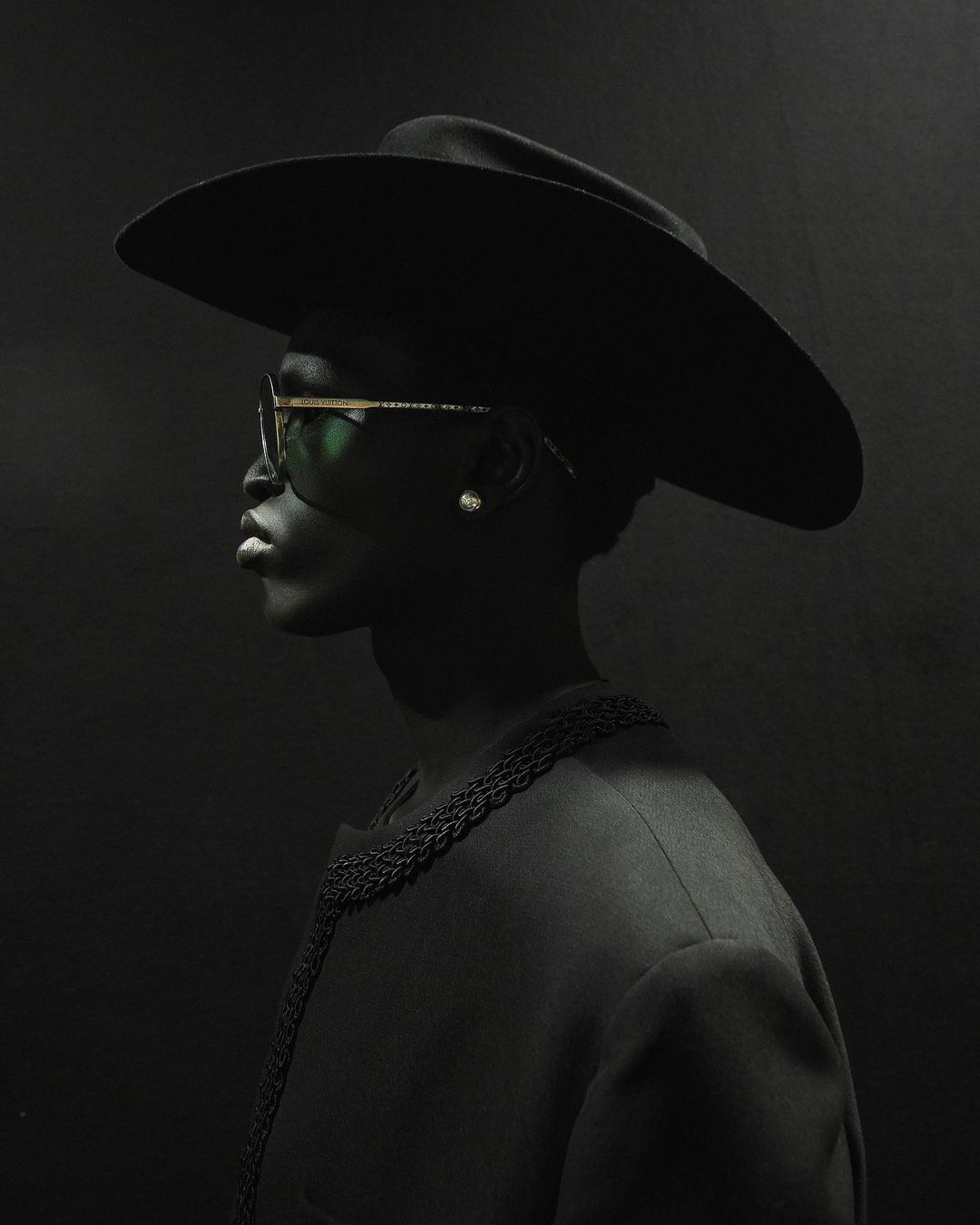 Profile portrait of a Black adult female wearing a wide-brimmed hat, sunglasses, and a jacket against a dark gray background.