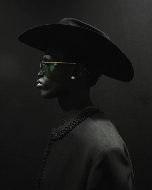 Profile portrait of a Black adult female wearing a wide-brimmed hat, sunglasses, and a jacket against a dark gray background.