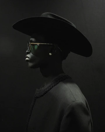 Profile portrait of a Black adult female wearing a wide-brimmed hat, sunglasses, and a jacket against a dark gray background.