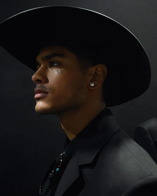 Portrait of a young adult man wearing a black cowboy hat and suit, side profile with a dark backdrop, illuminated for dramatic effect.