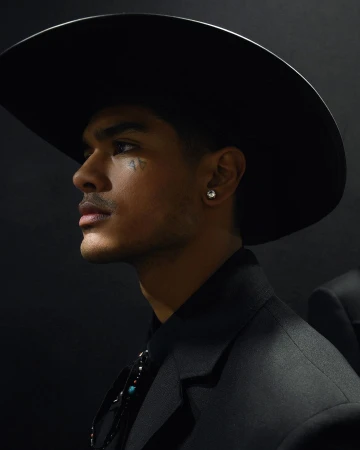 Portrait of a young adult man wearing a black cowboy hat and suit, side profile with a dark backdrop, illuminated for dramatic effect.