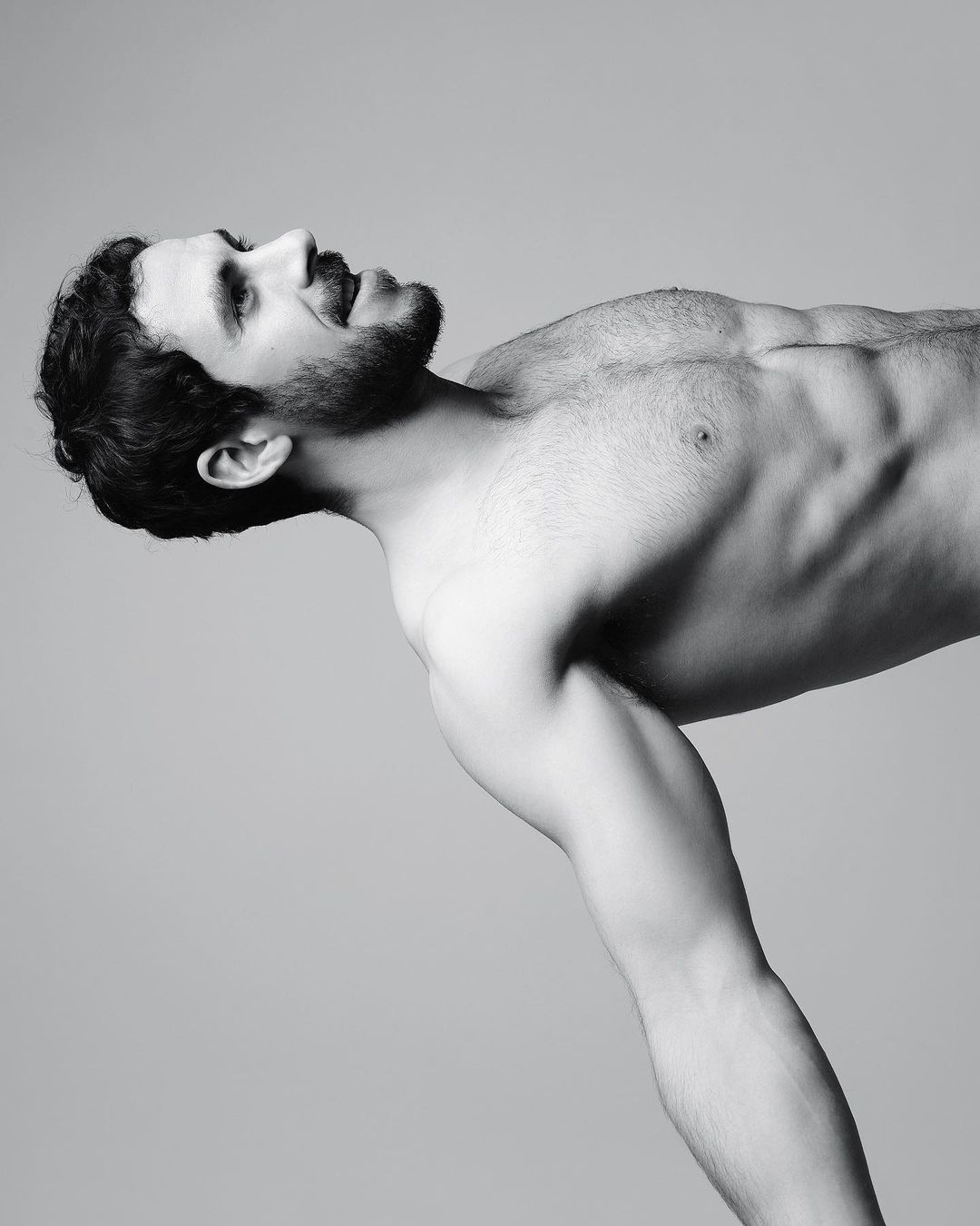Portrait of a shirtless man with a beard leaning forward against a plain grey studio backdrop