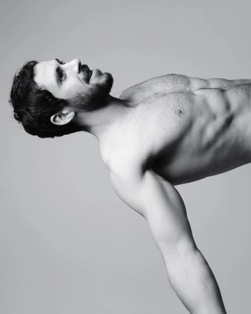 Portrait of a shirtless man with a beard leaning forward against a plain grey studio backdrop