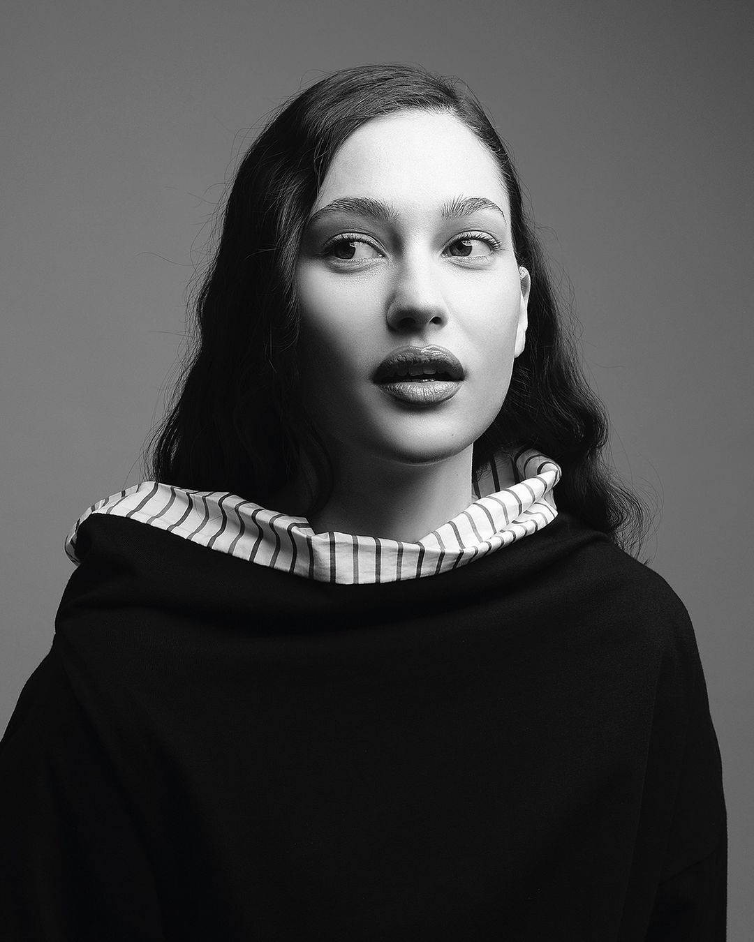 Portrait of a brunette adult female looking off-camera in black and white with soft studio lighting and dark clothing with striped trim.