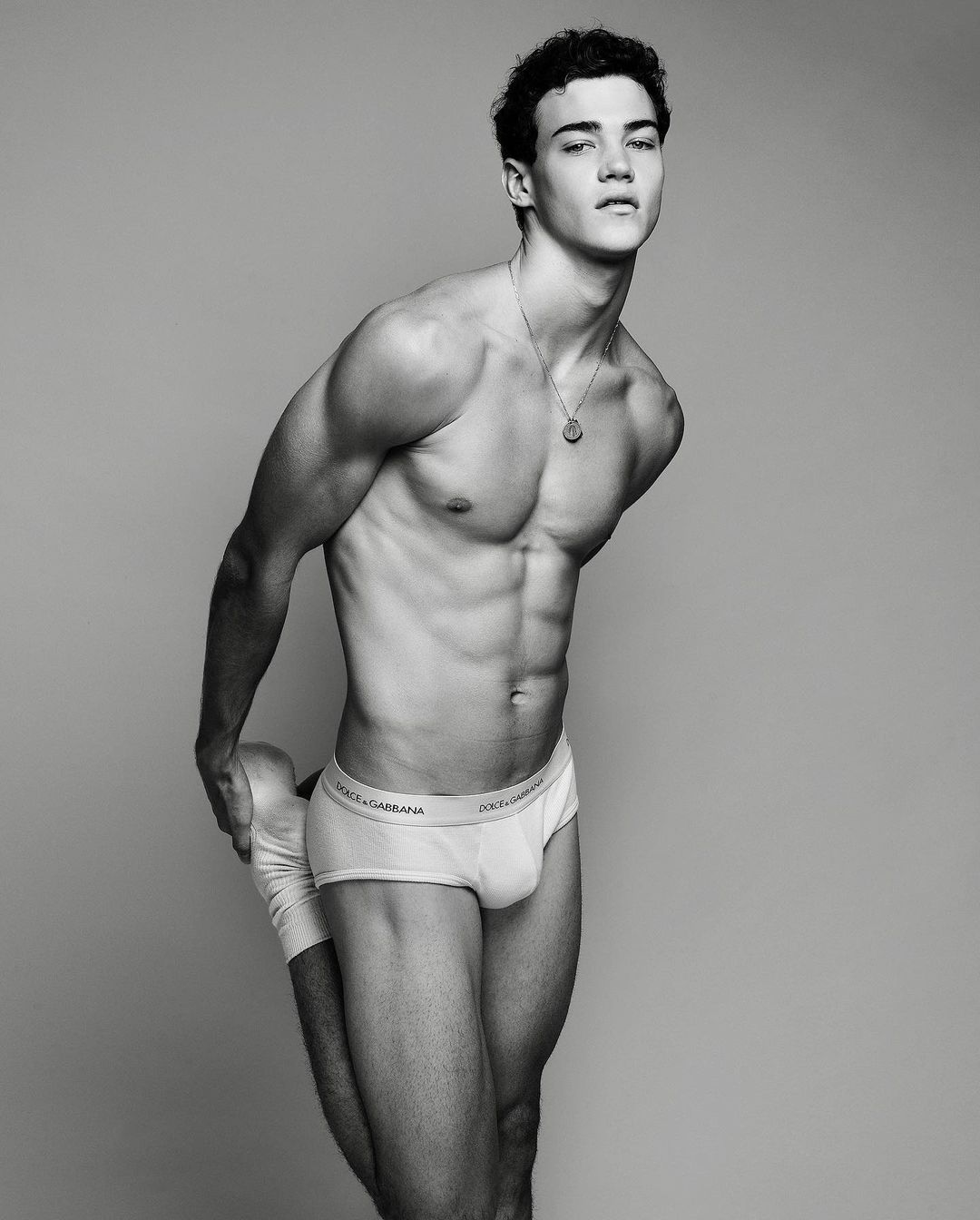 Shirtless athletic man in underwear stretches leg in a studio shot, showcasing his muscular physique against a plain background.