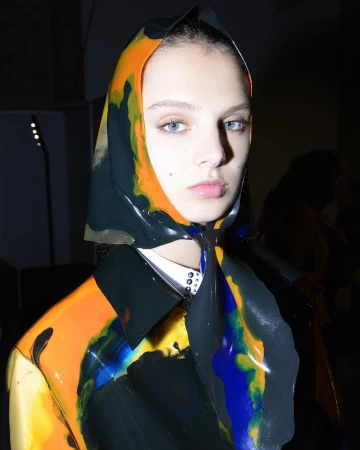 Close-up shot of a female fashion model wearing a patterned headscarf and high-shine vinyl jacket in a dimly lit indoor setting.